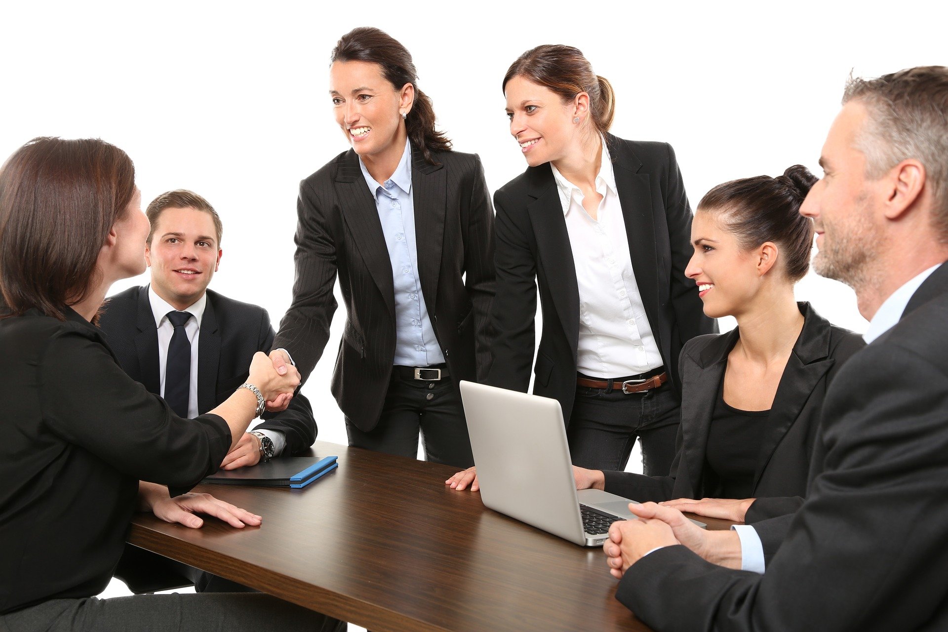 Eine Gruppe von 6 Menschen in Business-Kleidung reden miteinander. Eine Frau gibt einer anderen Frau über den Tisch die Hand. Insgesamt zwei Männer und zwei Frauen sitzen am Tisch. Zwei Frauen stehen am Tisch.